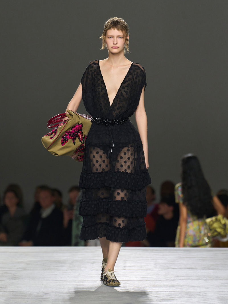 Model wearing a black lace dress on a runway with a blurred audience in the background