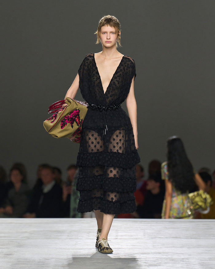 Model wearing a black lace dress on a runway with a blurred audience in the background