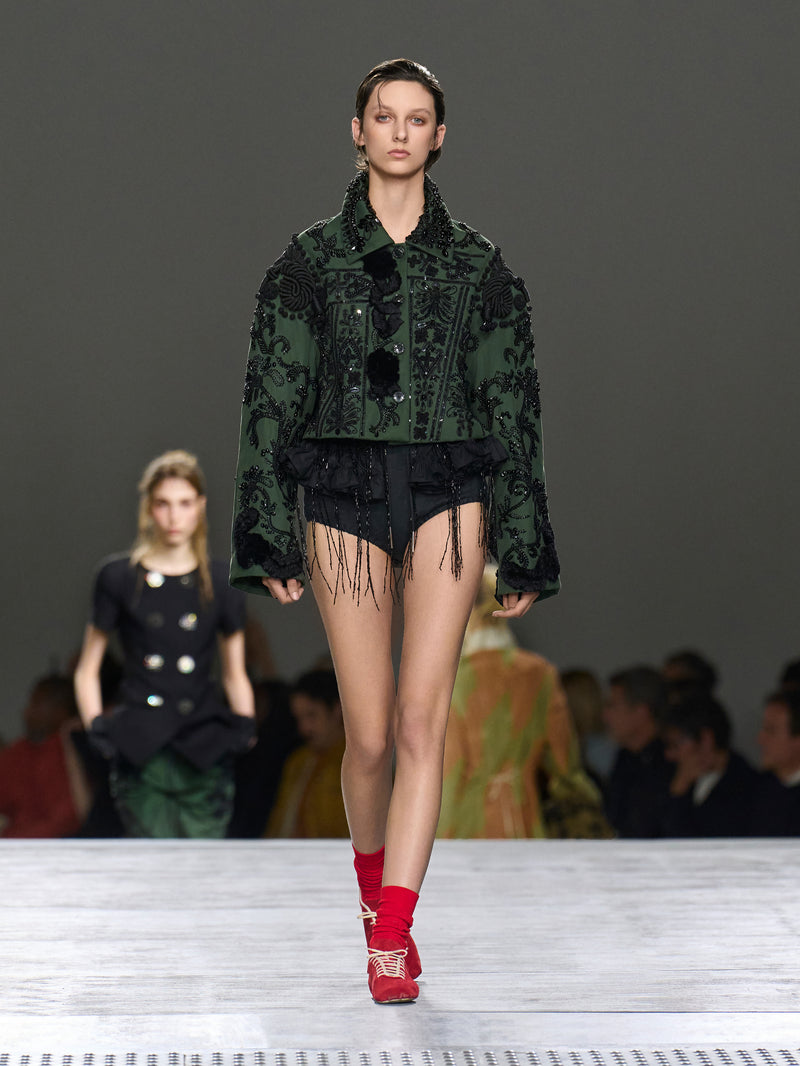 Model walking on a runway wearing a green patterned jacket and red shoes.
