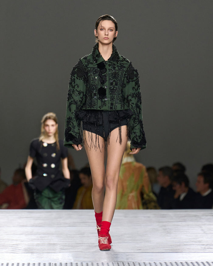 Model walking on a runway wearing a green patterned jacket and red shoes.