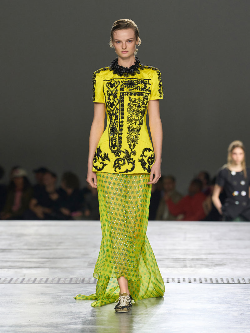 Model wearing a bright yellow and black dress on a runway with an audience in the background.
