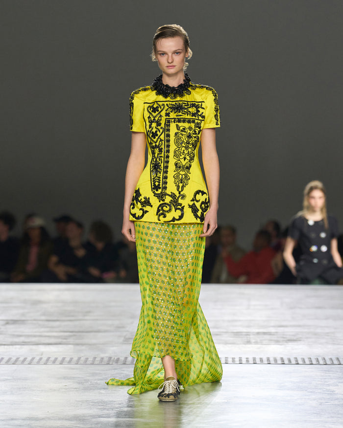 Model wearing a bright yellow and black dress on a runway with an audience in the background.