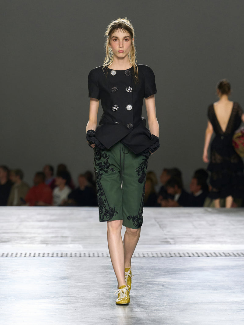 Model walking on a runway wearing a black top and green pants with floral embroidery.