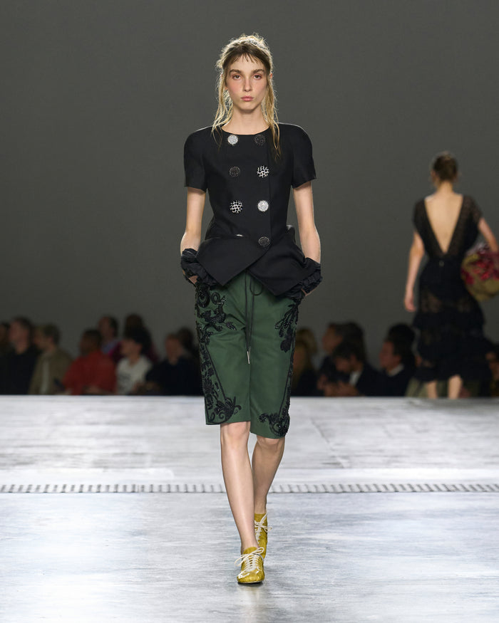 Model walking on a runway wearing a black top and green pants with floral embroidery.