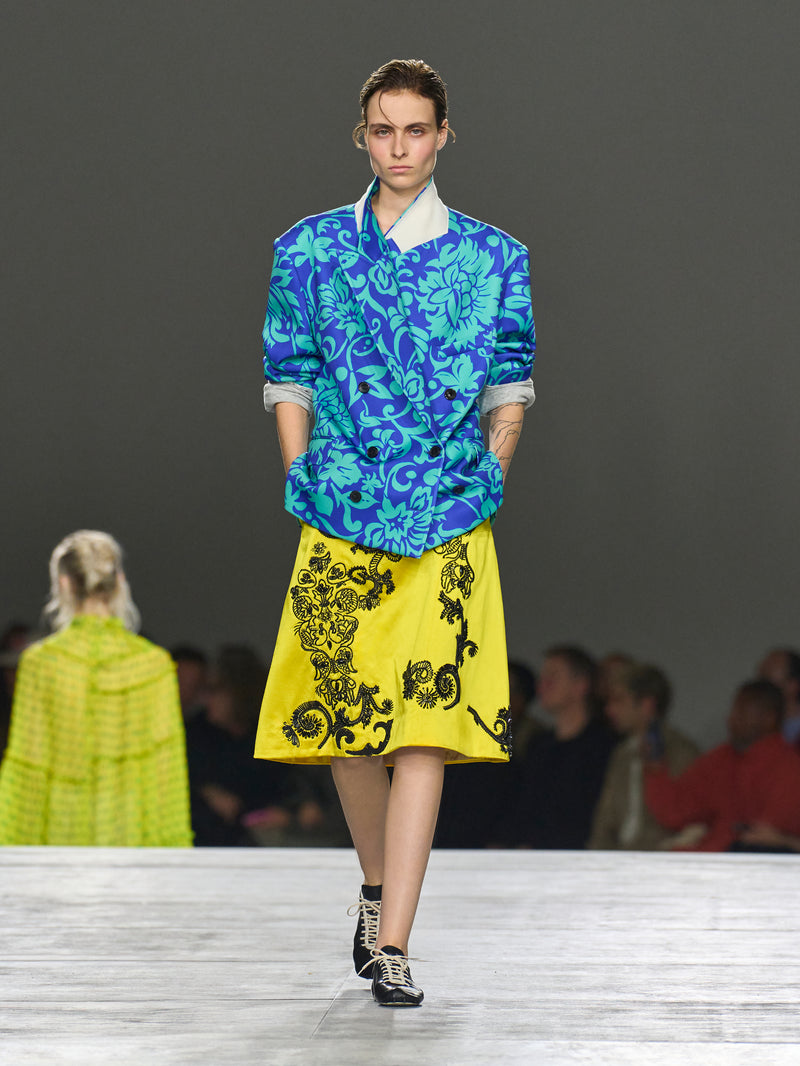 Model wearing a blue floral top and yellow skirt with black patterns on a runway.