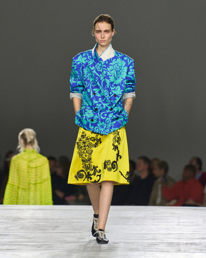 Model wearing a blue floral top and yellow skirt with black patterns on a runway.