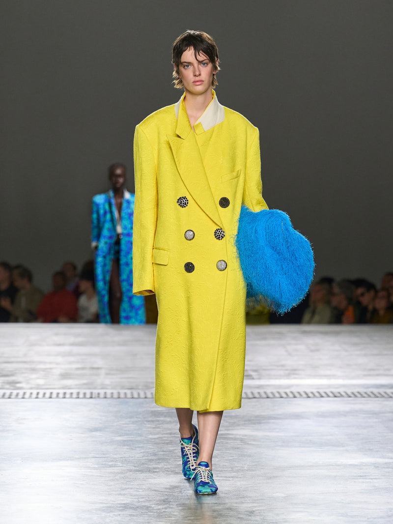Model wearing a bright yellow coat with a blue bag on a runway.
