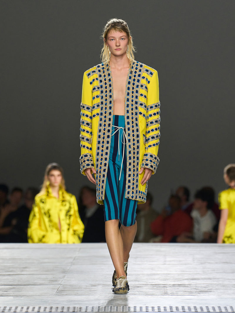Model walking on a runway wearing a yellow and patterned outfit with blue shorts.