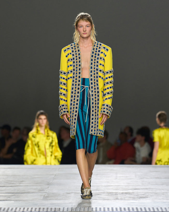 Model walking on a runway wearing a yellow and patterned outfit with blue shorts.