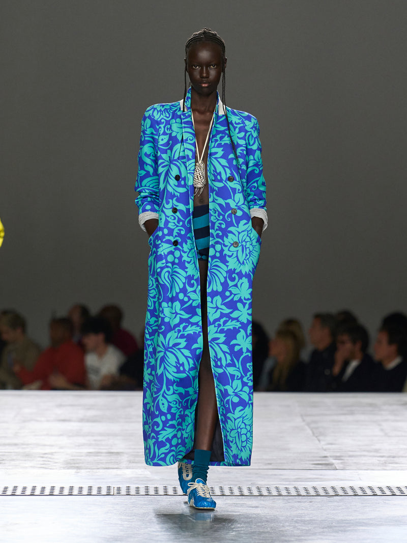 Model wearing a blue floral coat on a runway with an audience.