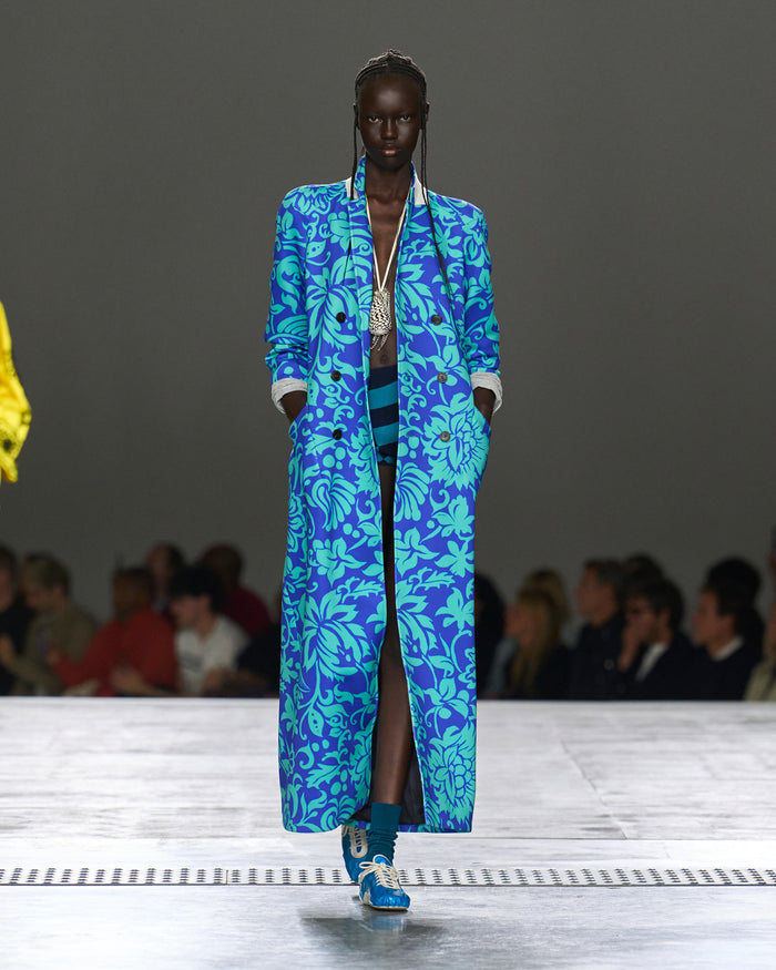 Model wearing a blue floral coat on a runway with an audience.