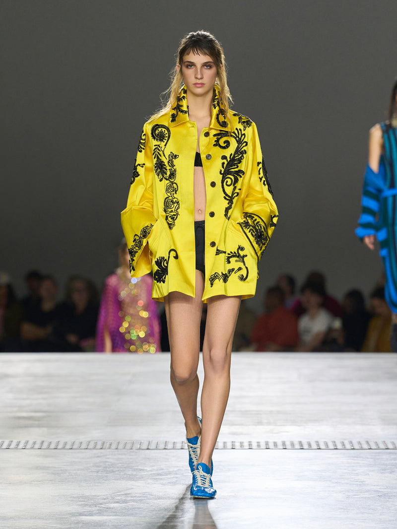Model wearing a bright yellow coat with black patterns on a runway.