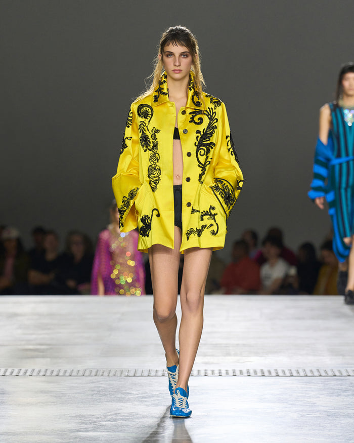 Model wearing a bright yellow coat with black patterns on a runway.