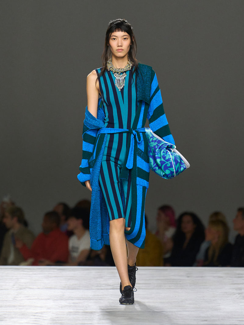 Model wearing a blue and green striped outfit with a patterned bag on a runway.