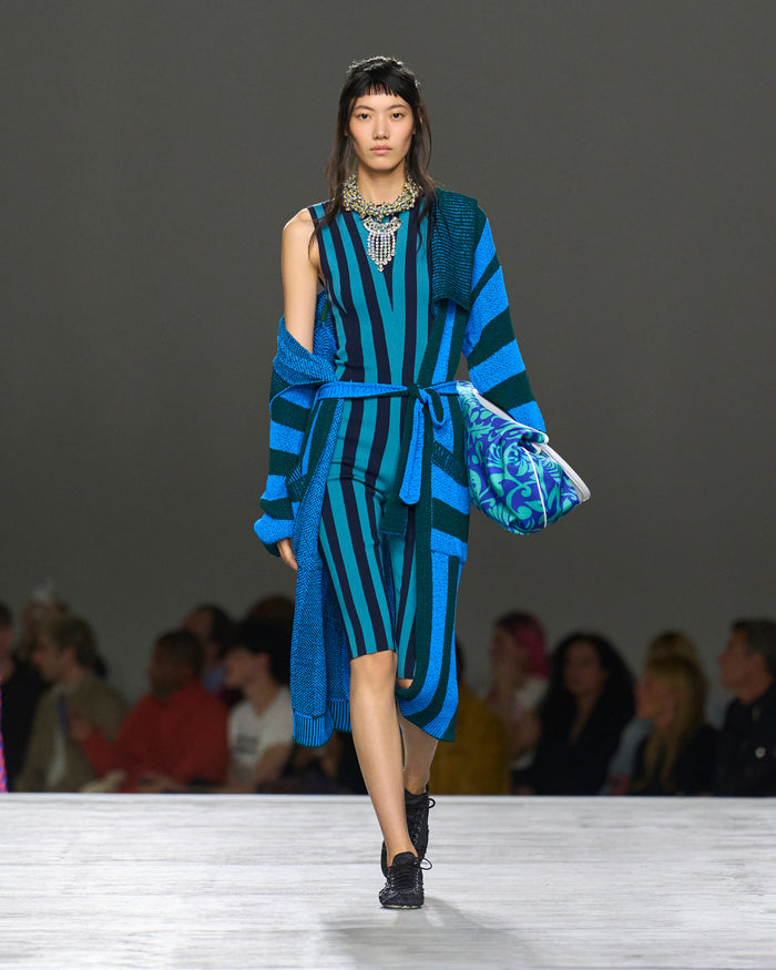 Model wearing a blue and green striped outfit with a patterned bag on a runway.