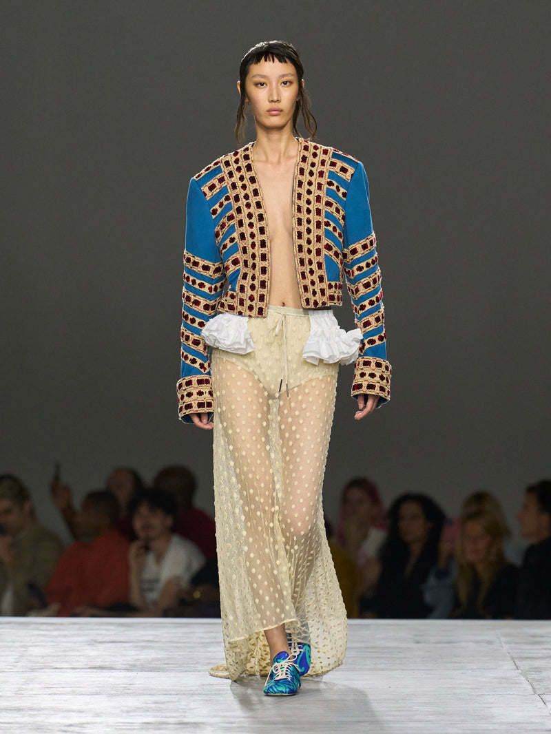 Model walking on a runway wearing a colorful outfit with a patterned jacket and sheer pants.