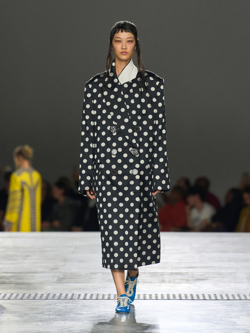 Model wearing a black coat with white polka dots on a runway.