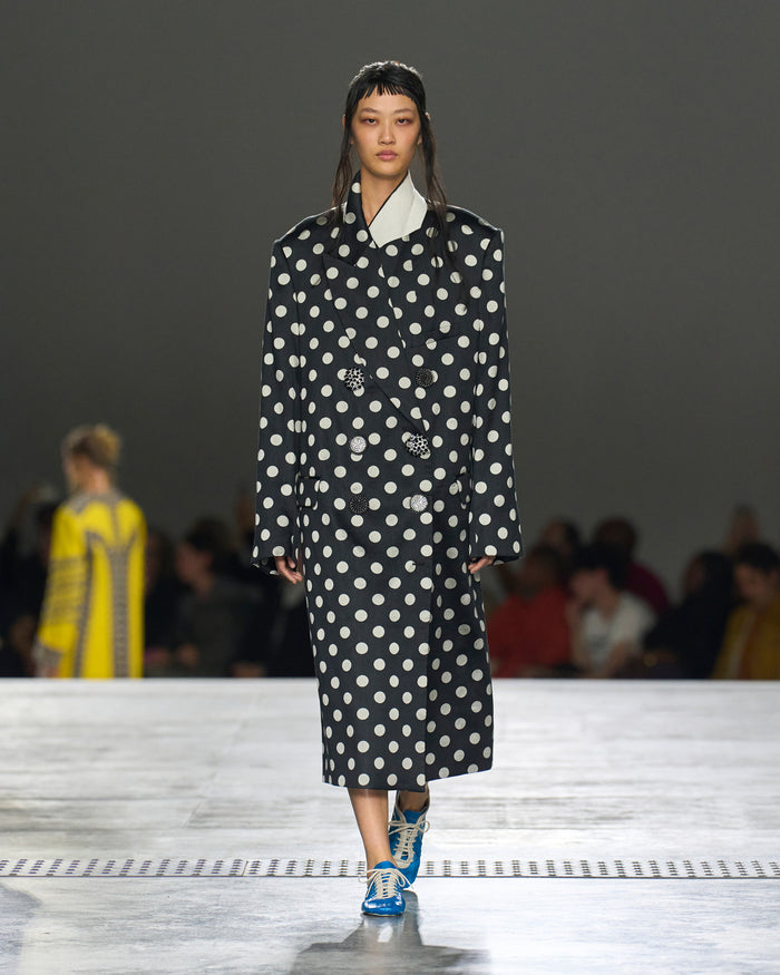 Model wearing a black coat with white polka dots on a runway.