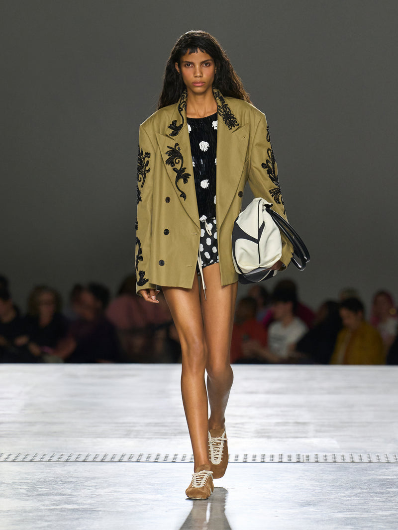 Model walking on a runway wearing a beige blazer with black floral embroidery, black and white polka dot dress, and white sneakers.