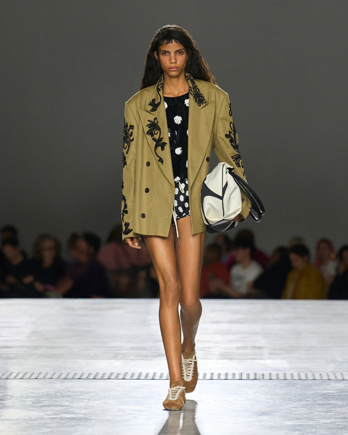 Model walking on a runway wearing a beige blazer with black floral embroidery, black and white polka dot dress, and white sneakers.