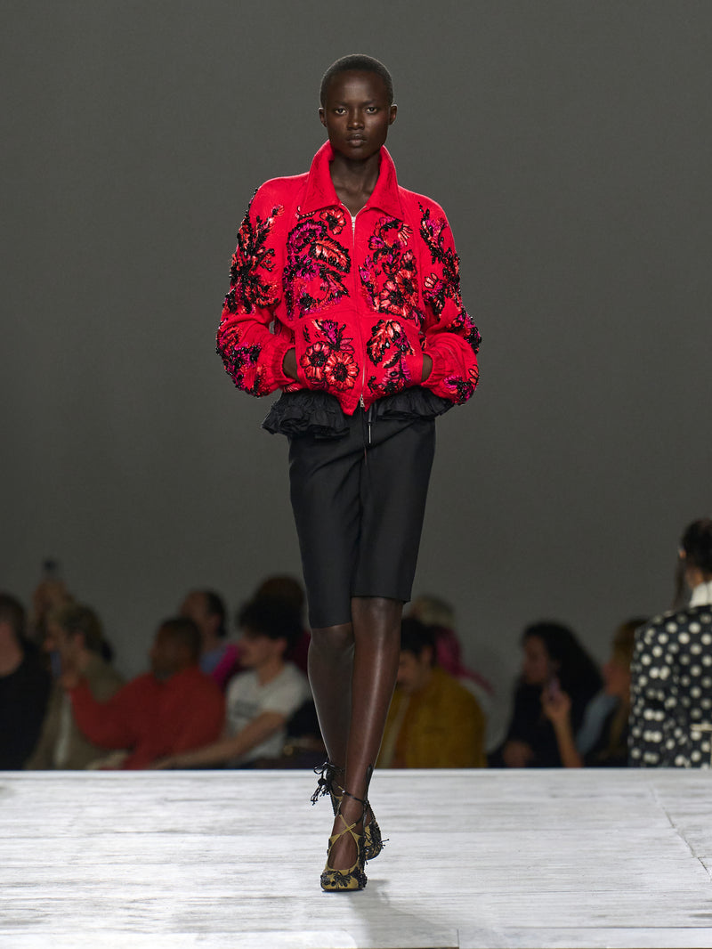Model wearing a red embroidered jacket on a runway with an audience.