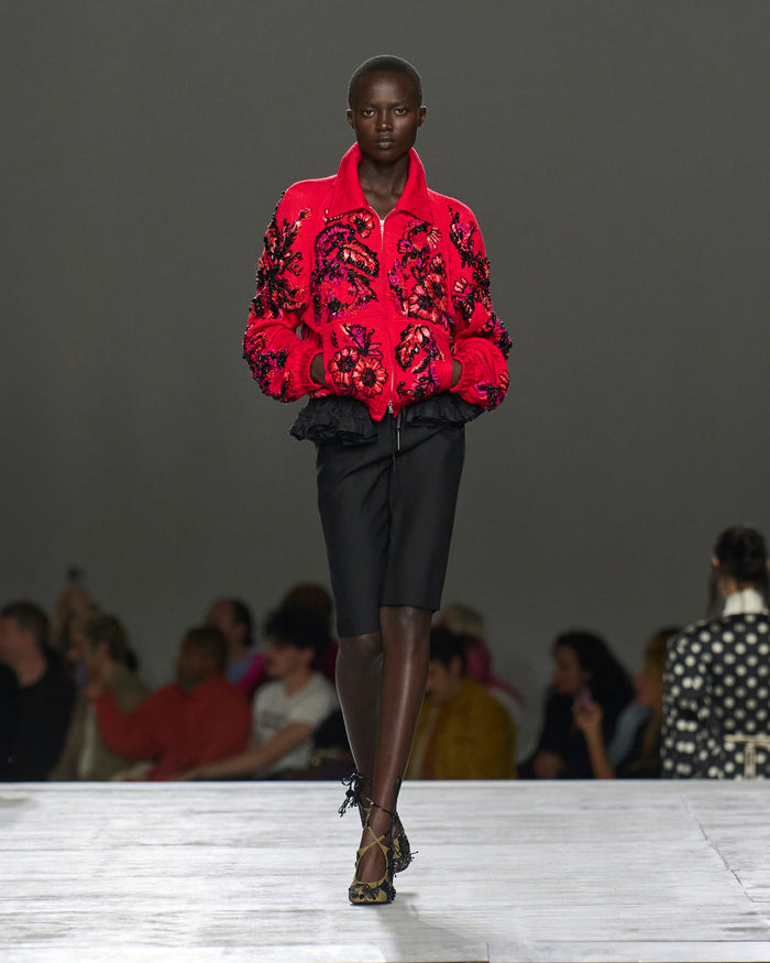 Model wearing a red embroidered jacket on a runway with an audience.