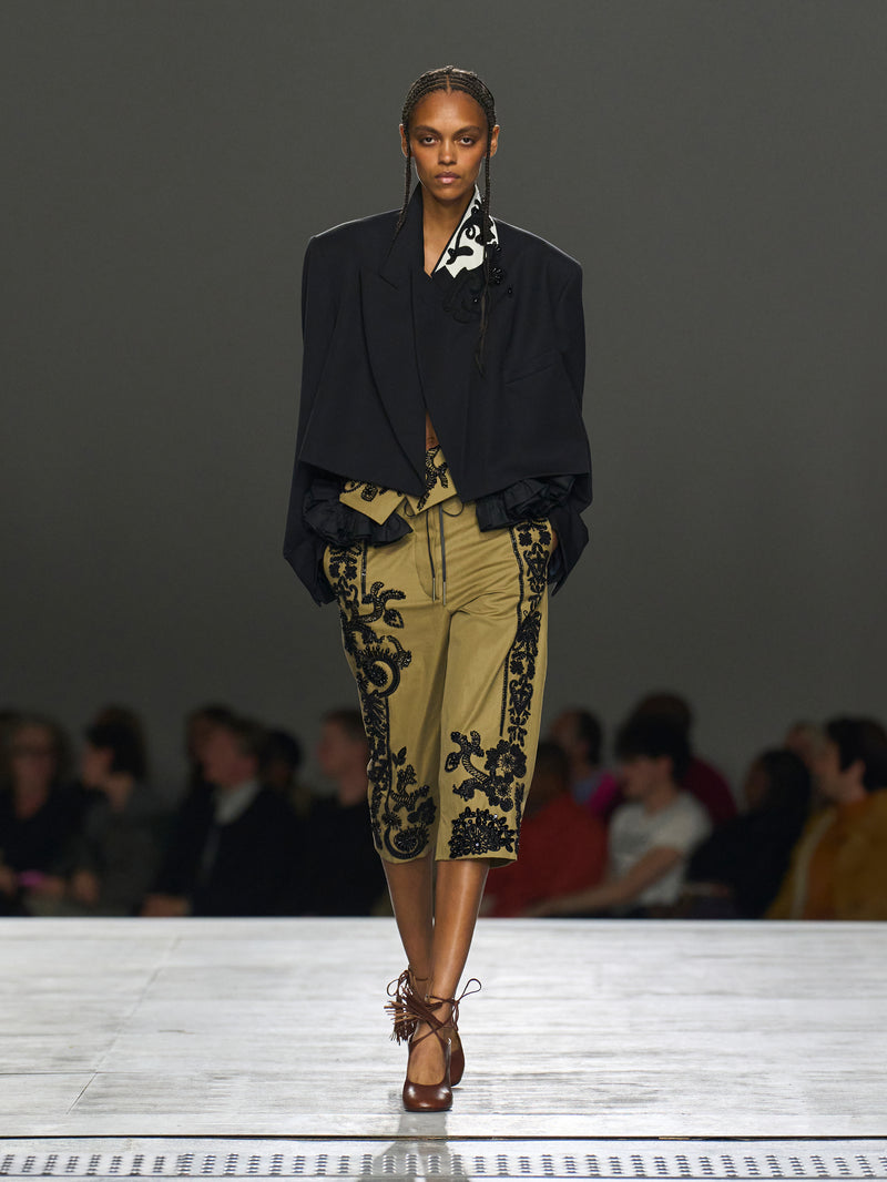 Model walking on a runway wearing a black blazer and patterned pants.