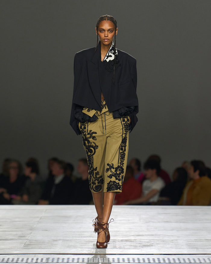 Model walking on a runway wearing a black blazer and patterned pants.