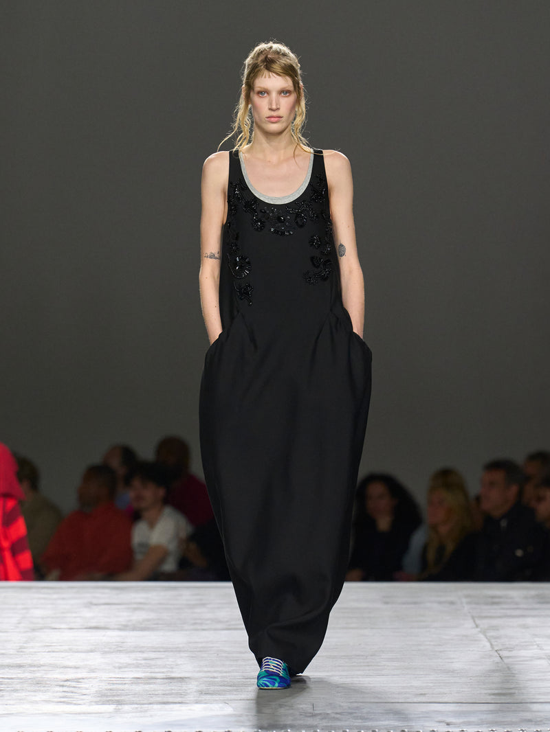 Model walking on a runway wearing a black dress with embellishments.