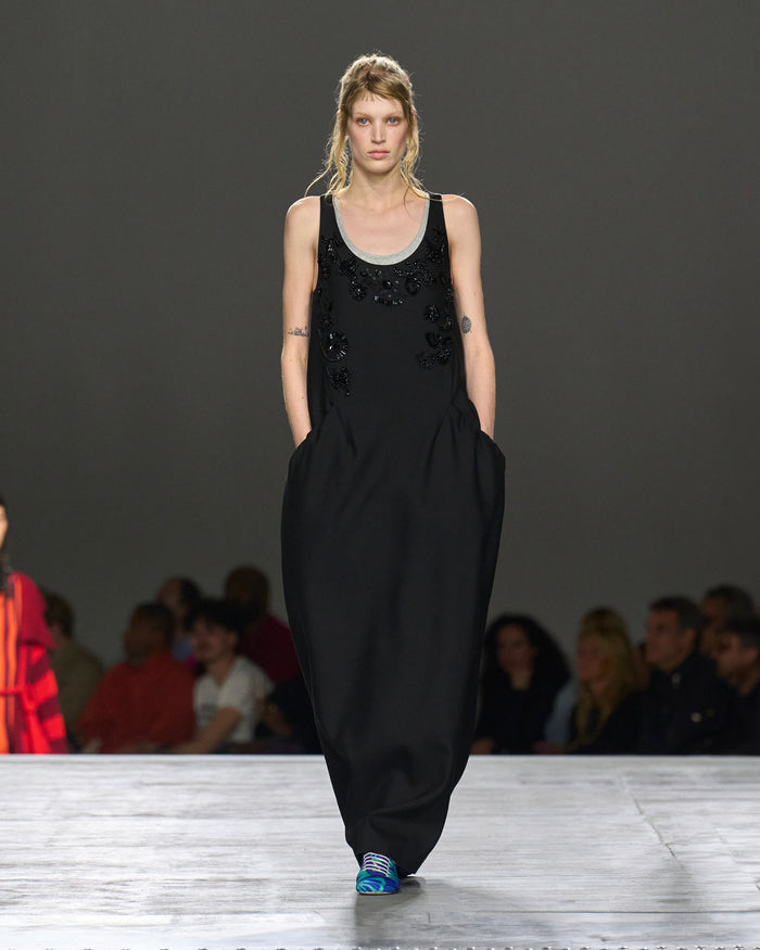 Model walking on a runway wearing a black dress with embellishments.
