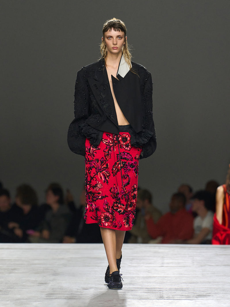 Model walking on a runway wearing a black jacket and red floral skirt.