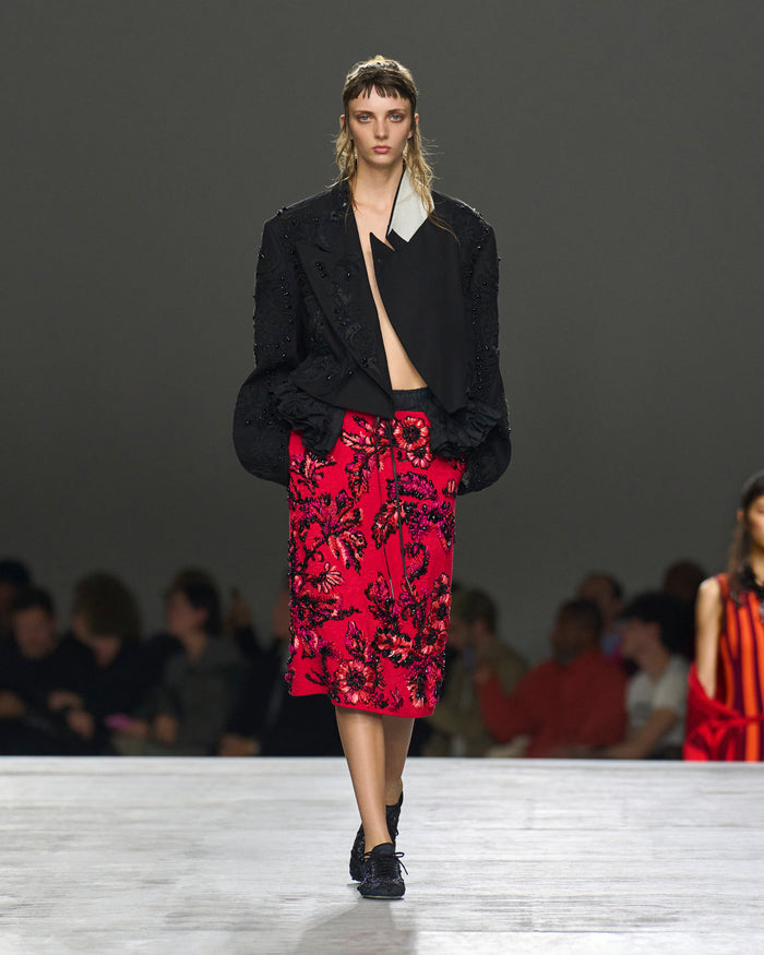 Model walking on a runway wearing a black jacket and red floral skirt.