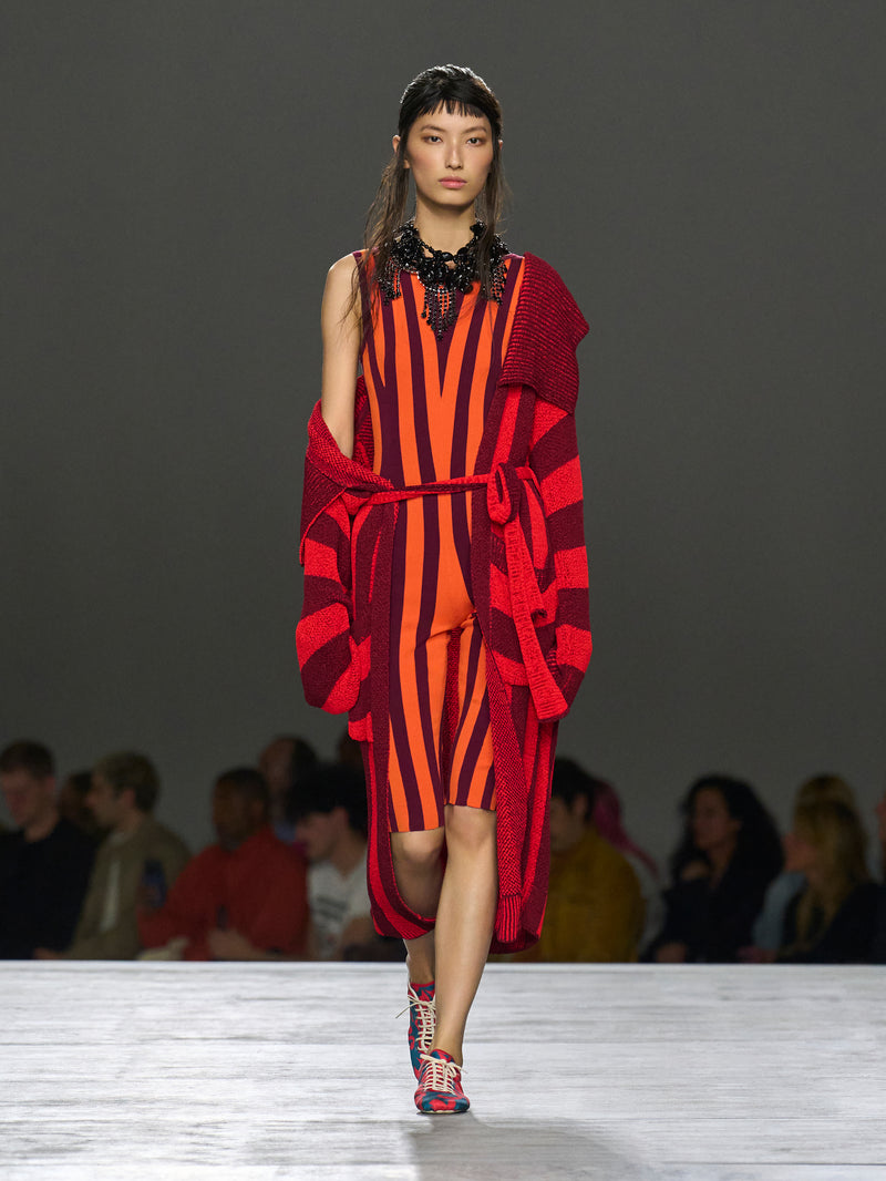 Model walking on a runway wearing a red and orange striped dress with a matching cardigan.