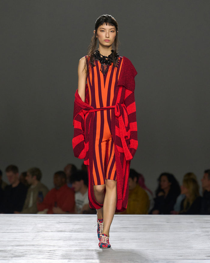 Model walking on a runway wearing a red and orange striped dress with a matching cardigan.