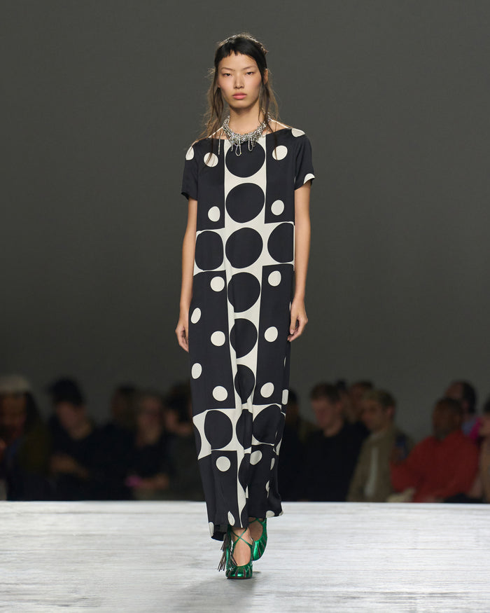 Model wearing a black and white polka dot dress on a runway with an audience.