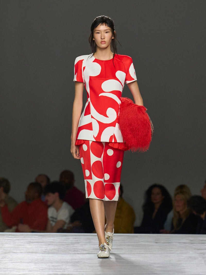 Model wearing a red and white outfit with a large red bag on a runway.