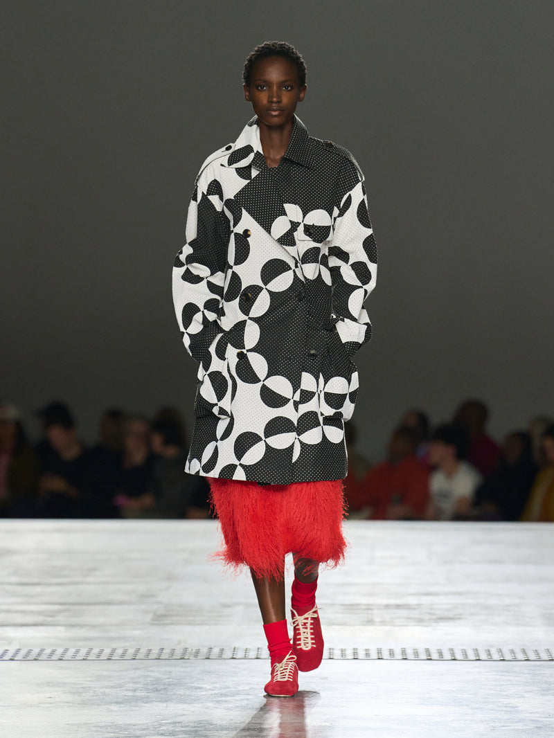 Model walking on a runway wearing a black and white patterned coat with a red skirt.
