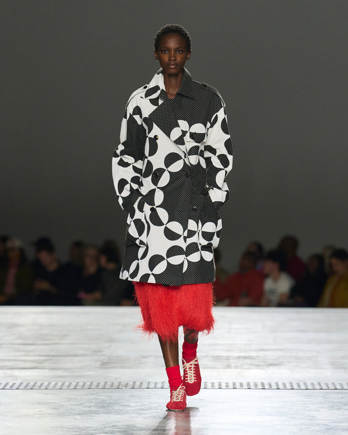 Model walking on a runway wearing a black and white patterned coat with a red skirt.