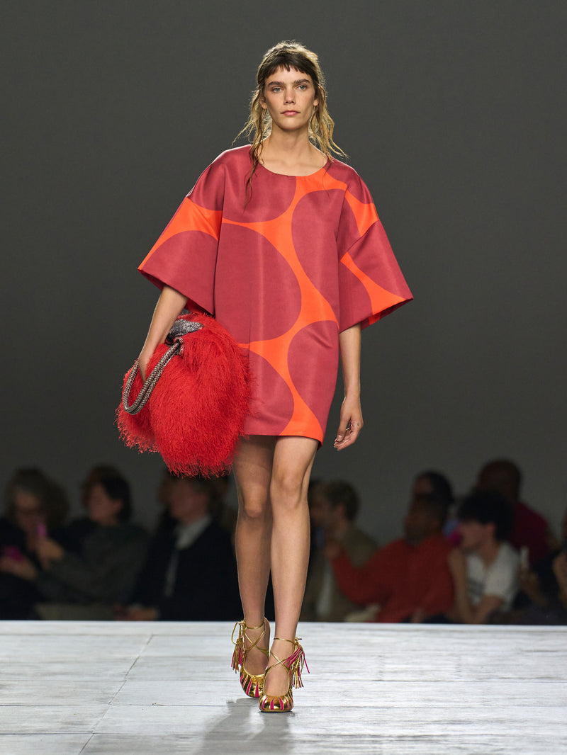 Model wearing a red and orange dress with a feathered handbag on a runway.