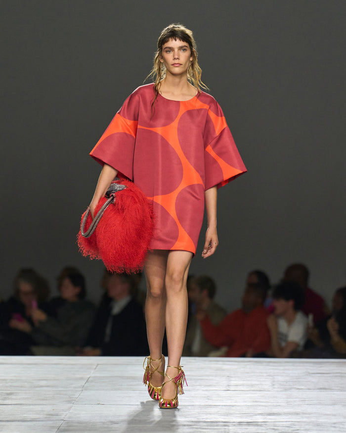 Model wearing a red and orange dress with a feathered handbag on a runway.
