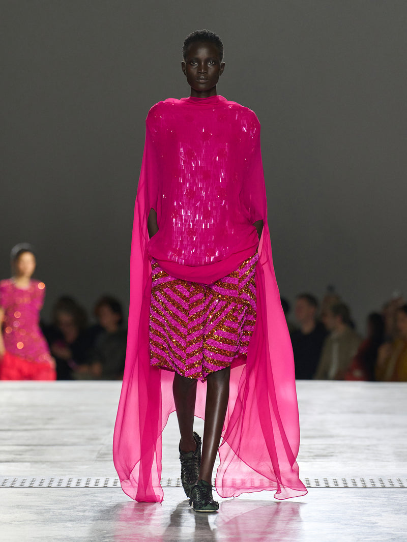 Model wearing a bright pink outfit with a sheer overlay on a runway.