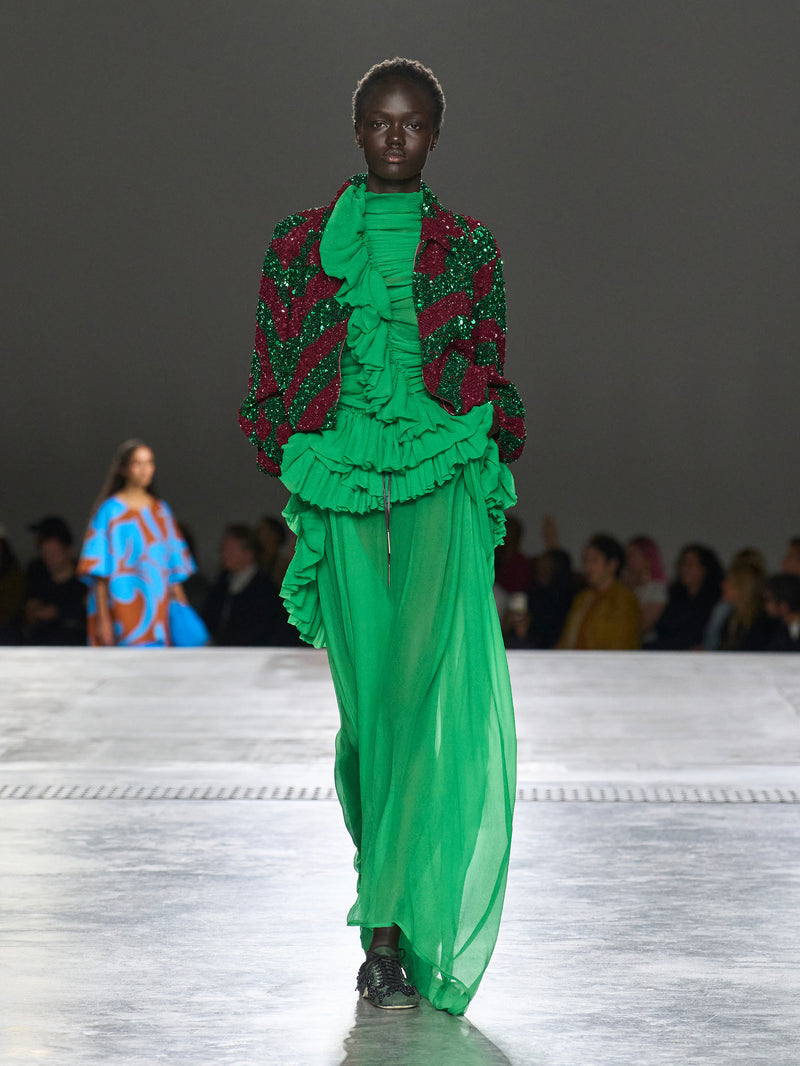 Model wearing a vibrant green and red outfit on a runway with an audience.