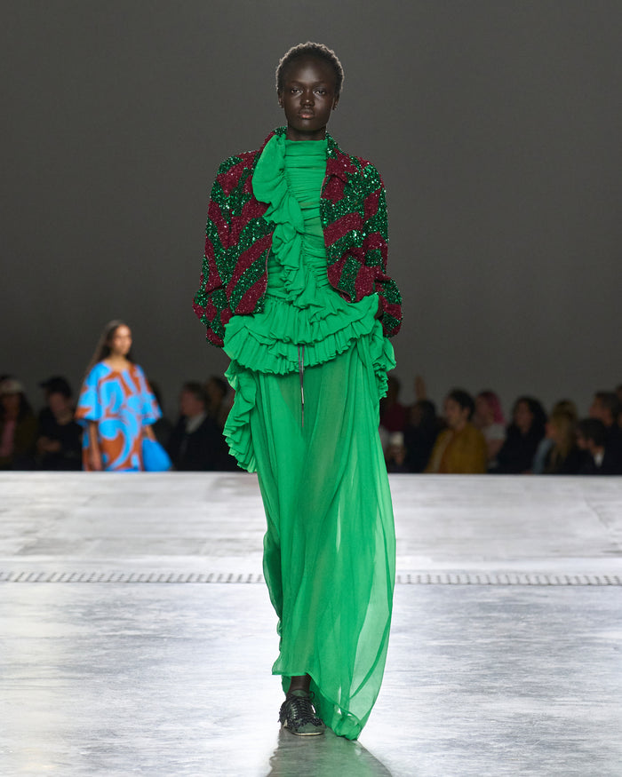 Model wearing a vibrant green and red outfit on a runway with an audience.