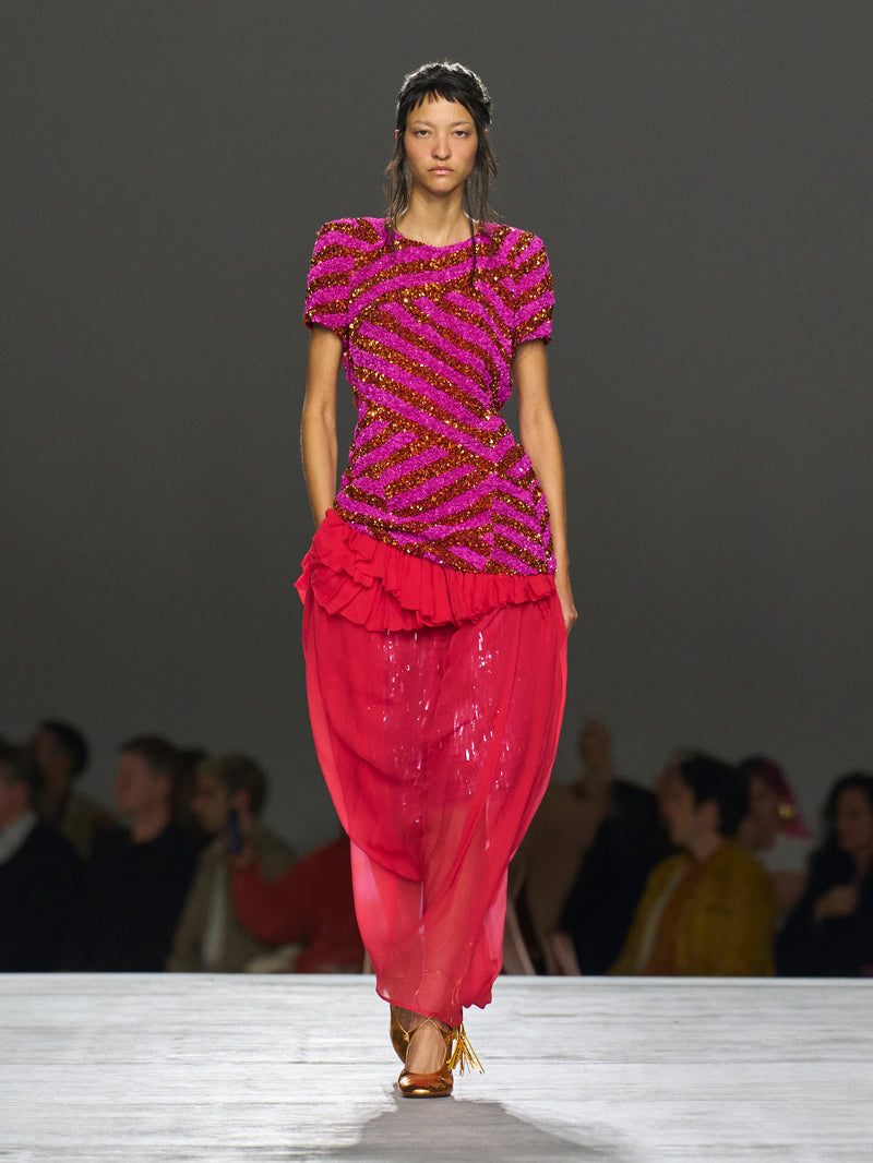 Model wearing a vibrant pink and purple outfit on a runway with an audience.