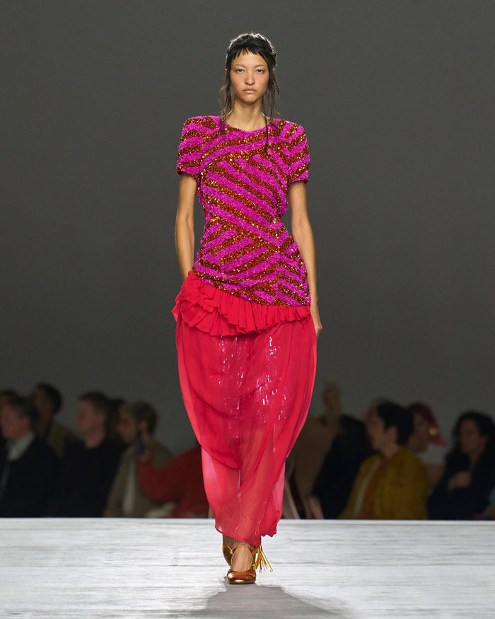 Model wearing a vibrant pink and purple outfit on a runway with an audience.
