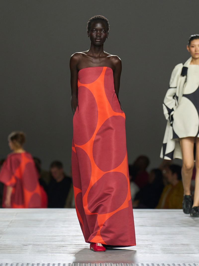 Model wearing a red and orange patterned dress on a fashion runway.