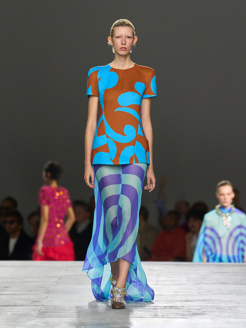 Model walking on a runway wearing a colorful outfit with abstract patterns.