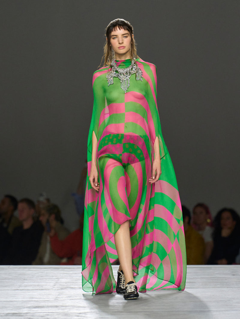 Model wearing a vibrant pink and green patterned dress on a runway with an audience.