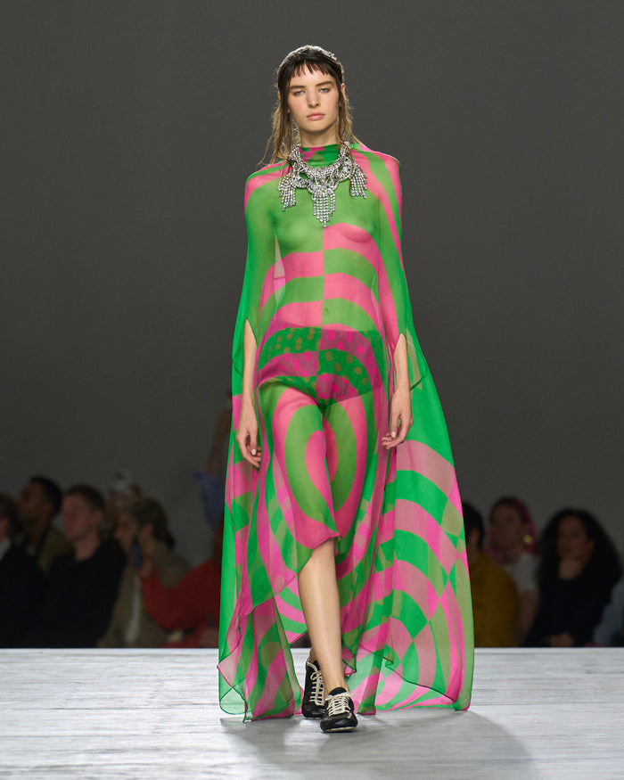 Model wearing a vibrant pink and green patterned dress on a runway with an audience.