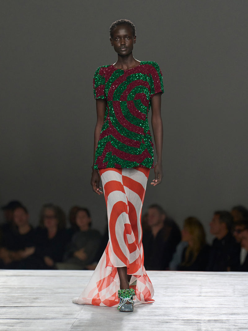 Model wearing a vibrant, patterned outfit on a runway with an audience in the background.